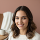 Headshot of a smiling young woman with natural makeup and soft lighting against a blurred warm brown and dusty rose backgroun